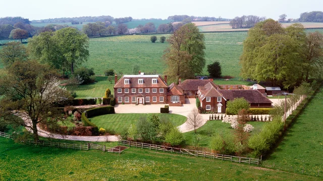 Elevated view of a red-brick country house, outbuildings and a curved drive, with lawns and fields beyond.