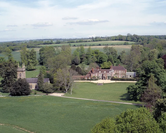 Elevated view across open grass towards a country house and neighbouring buildings among mature trees.