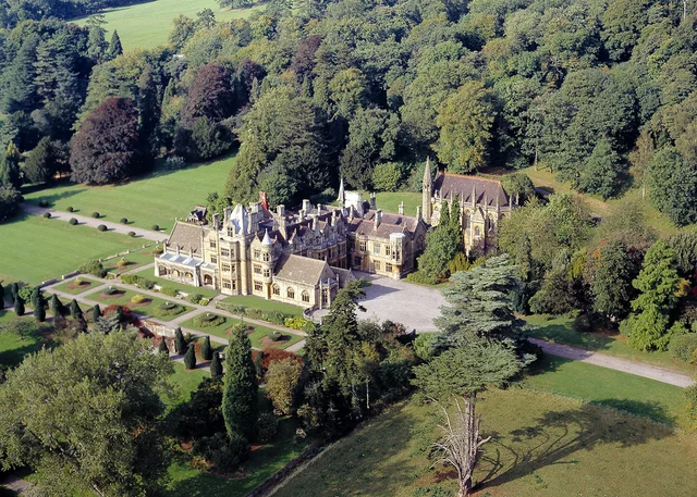 Elevated view of a large stone house with towers and formal gardens, bordered by woodland and lawns.