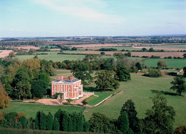 Elevated view of a red-brick house with white window surrounds, set among lawns, trees and open fields.