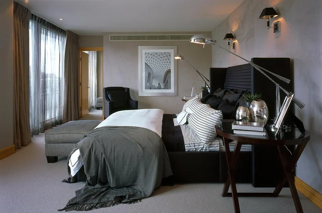 A contemporary bedroom with dark bedding, a tall upholstered headboard and daylight filtering through sheer curtains.