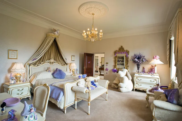 An ornate bedroom with a draped bed canopy, pale painted furniture, purple accents and a chandelier.