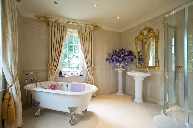 A white freestanding bath beneath a curtained window, beside a pedestal basin, gilded mirror and glass shower enclosure.