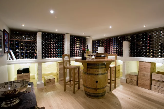 Wine racks line a softly lit cellar around a central barrel table and stools, with wooden boxes along the floor.