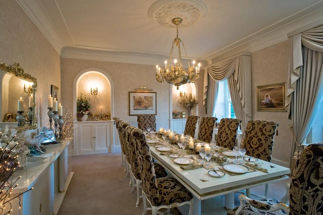 A formal dining room with a set table, patterned upholstered chairs, a chandelier and tall curtained windows.