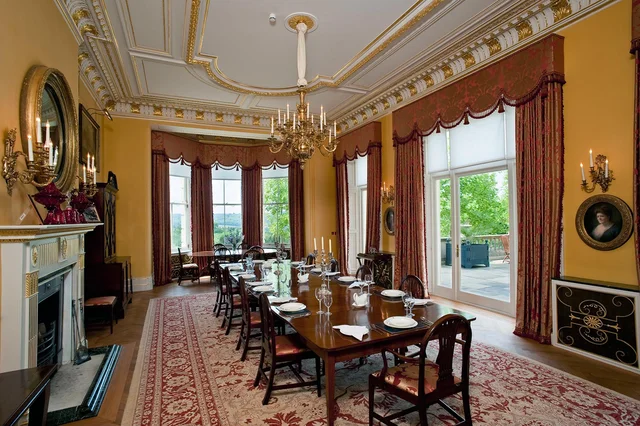 A long dining table stands in a room with golden walls, red curtains, an ornate ceiling and a fireplace.