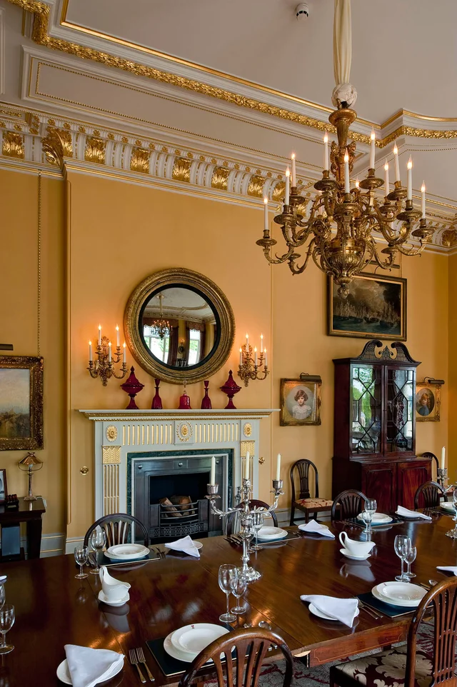 Detail of a dining room with a set wooden table, a fireplace, a round mirror and a chandelier against golden walls.