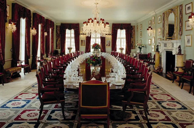 A long polished dining table surrounded by dark chairs in a grand room with red curtains, chandeliers and a patterned rug.