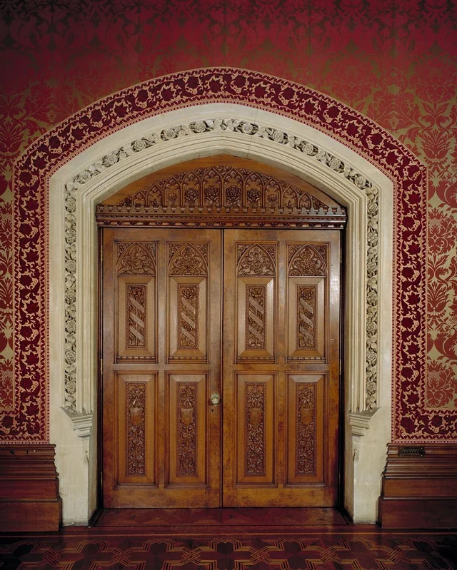 Carved wooden double doors sit within an elaborate pale arch against patterned red walls.