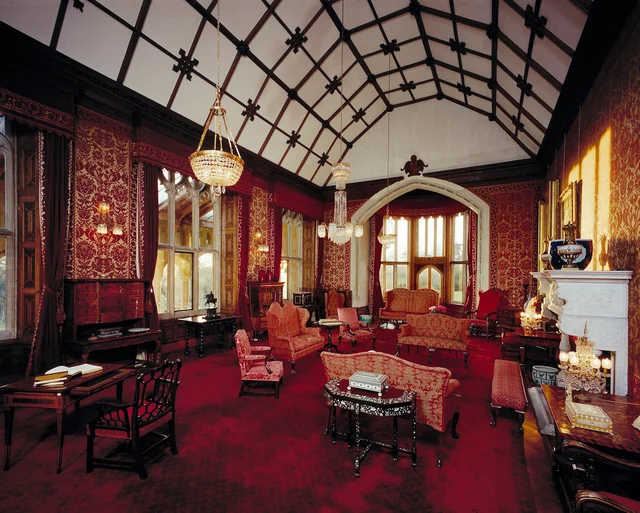 A red drawing room with dark wood panelling, upholstered seating, a fireplace and a high patterned vaulted ceiling.