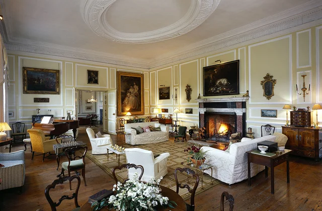 A spacious drawing room with pale wall panels, framed paintings, white seating and a lit fireplace.