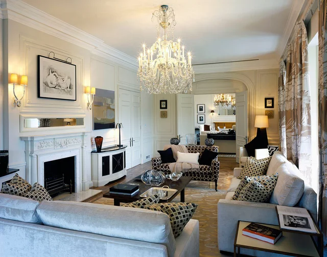 A pale sitting room with upholstered seating, a fireplace, patterned cushions and a crystal chandelier.