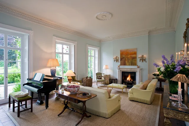A light drawing room with tall garden-facing windows, pale seating, a piano and a lit fireplace.