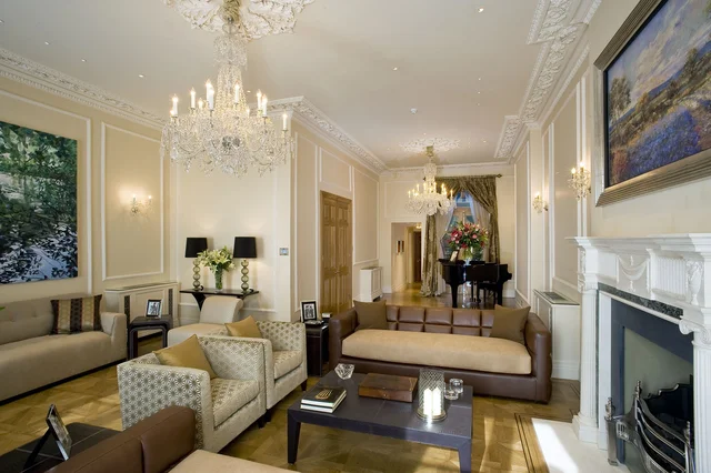 A long pale drawing room with sofas, a glass coffee table, two chandeliers and a piano at the far end.