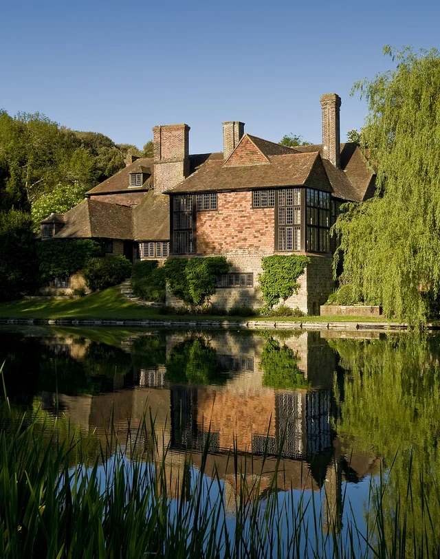 A brick-and-timber house beside still water, reflected below with reeds in the foreground and a willow to one side.