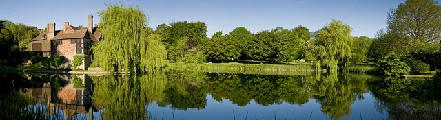A wide view of a house, weeping willow and surrounding trees reflected in a still stretch of water.