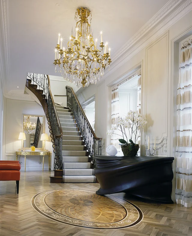 A bright entrance hall with a broad staircase, a grand piano, a large chandelier and a circular floor pattern.