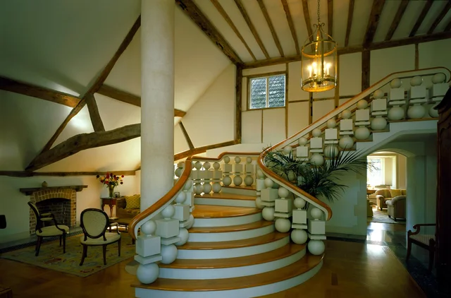 A sweeping staircase with rounded pale balusters rises through a hall with exposed timbers and a hanging lantern.