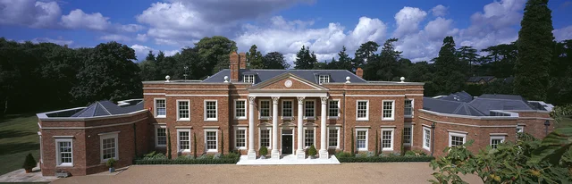 A symmetrical red-brick house with a tall columned entrance, lower side wings and a broad gravel forecourt.