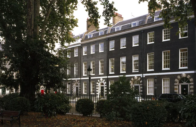 A tall dark-brick townhouse with rows of white-framed windows, seen from a shaded garden.