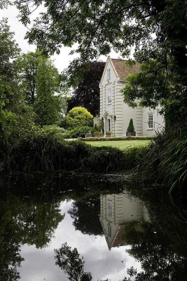 A pale gabled house partly framed by trees and reflected in a narrow stretch of water.
