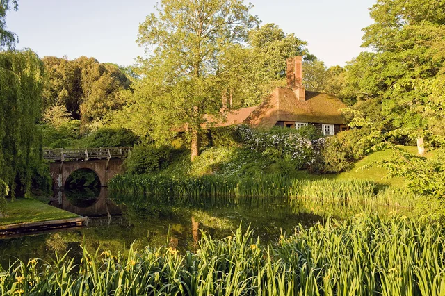 A low house with a tiled roof stands behind trees beside an arched bridge and water edged with reeds.