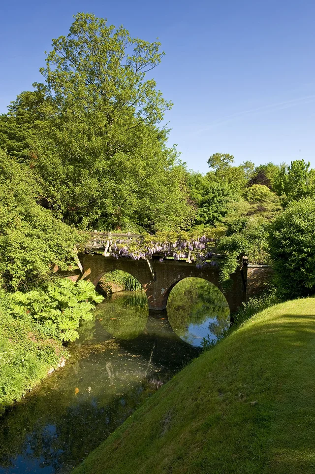 A small arched bridge crosses a narrow waterway between grassy banks and leafy trees.