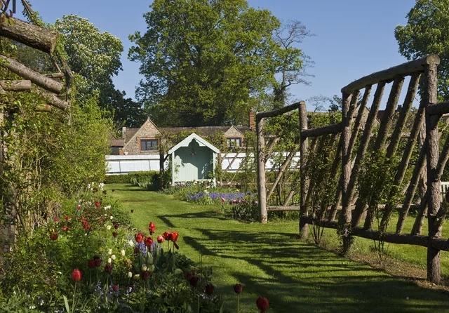 An open wooden gate leads along a grassy path beside flowering borders towards a small garden building.
