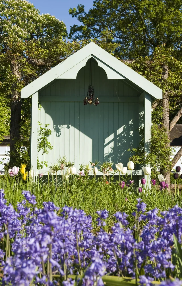 A small green garden shelter with white trim stands beyond a bed of purple flowers.