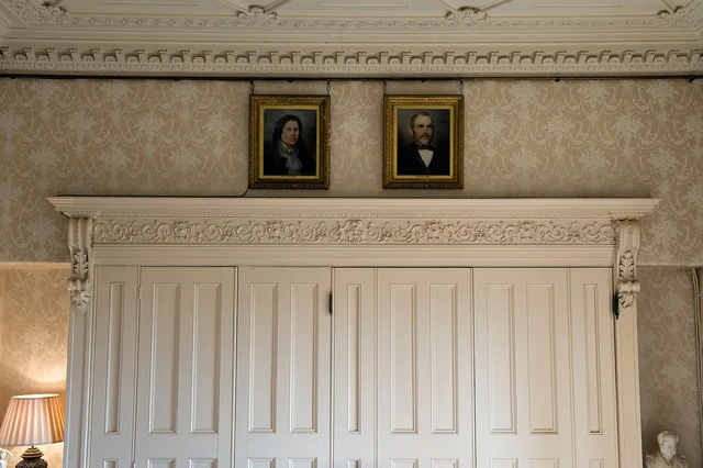Two small framed portraits hang above pale double doors with an elaborately carved surround.