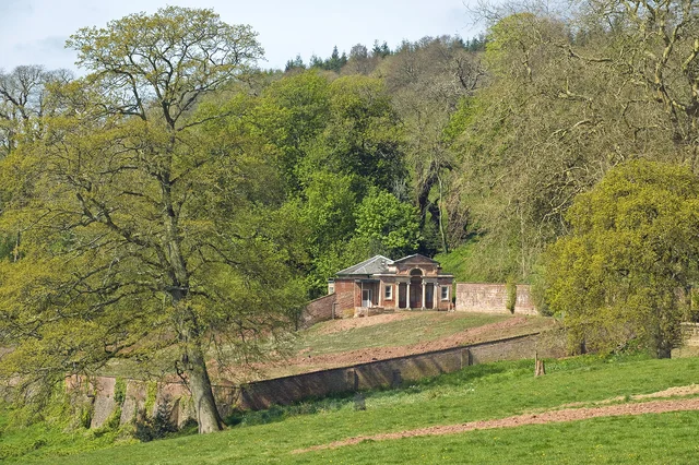 A small building stands beyond a brick wall in a green landscape of grass and mature trees.