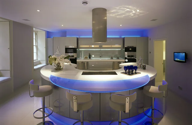 A contemporary kitchen centres on a curved white island with stools and blue accent lighting beneath a metal extractor.