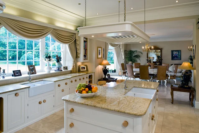 A pale kitchen with an island, overhead extractor and curtained garden-facing windows opens towards a sitting area.