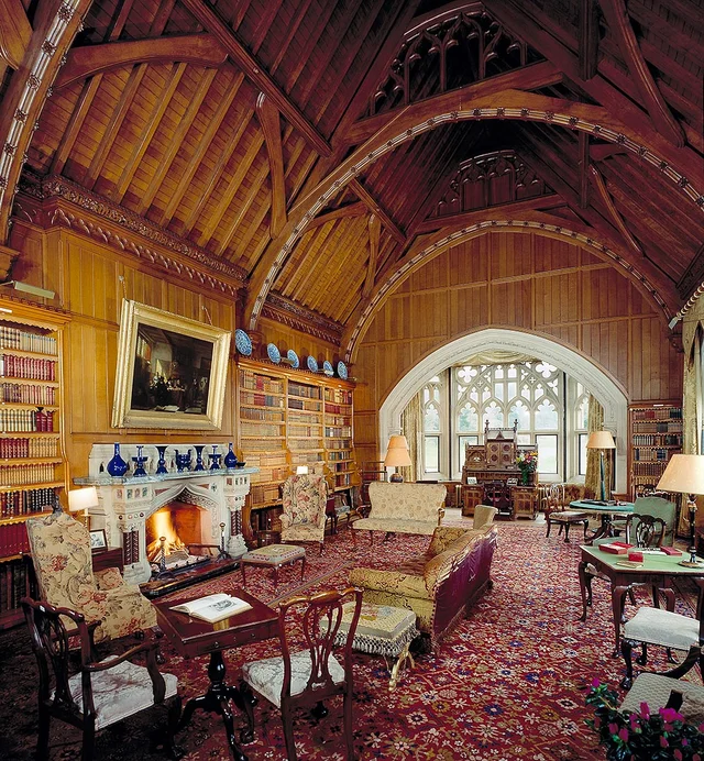 A library with dark timber walls, a high exposed roof, bookshelves and patterned seating faces a large arched window.