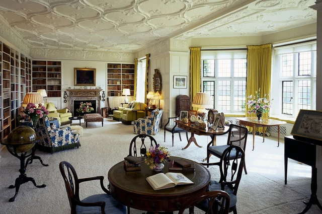 A spacious library with book-lined shelves, upholstered chairs, round tables and tall windows beneath an ornate pale ceiling.