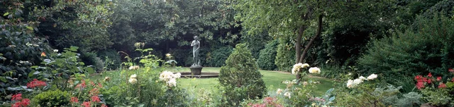 A garden statue stands beyond a lawn framed by flowering borders and dense trees.
