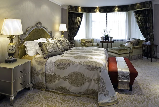 A patterned bedspread and upholstered bench sit beside a broad bay window with dark curtains.
