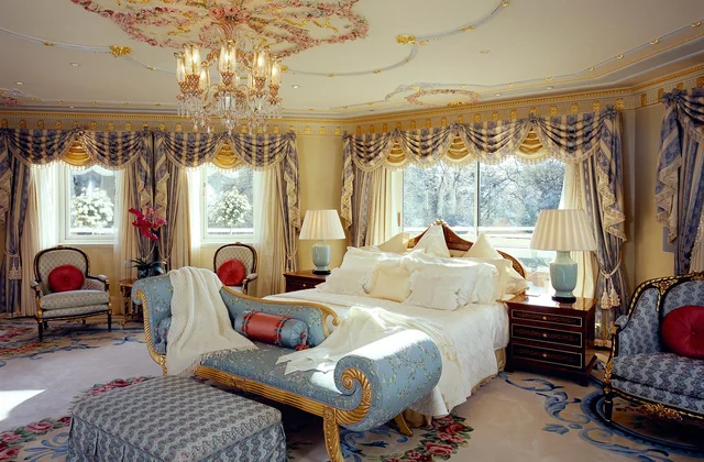 A chandelier hangs above a bedroom with elaborate drapery, a decorative ceiling and a pale blue chaise longue.