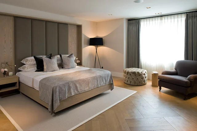 A bed with a tall panelled headboard sits beside a large curtained window and an armchair.