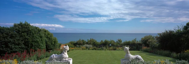 Two deer sculptures flank a lawn overlooking a broad expanse of water beneath a blue sky.