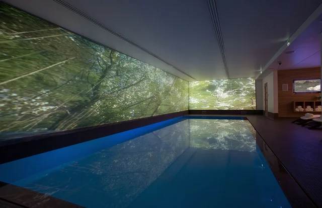 An indoor swimming pool reflects large woodland images covering two walls, with loungers and folded towels to one side.