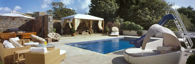 An outdoor swimming pool is surrounded by cushioned seating, a shaded pavilion and a slide.