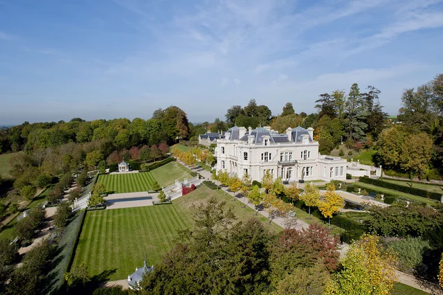 An elevated view of a large pale house surrounded by striped lawns, terraces and formal planting.