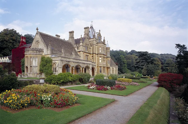 An ornate house with gables and tall chimneys rises behind colourful flower beds and intersecting paths.