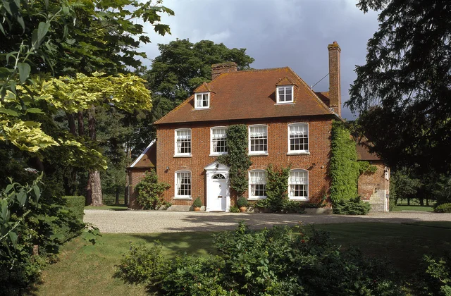 A red-brick house with a white entrance and dormer windows stands among trees and shaded planting.