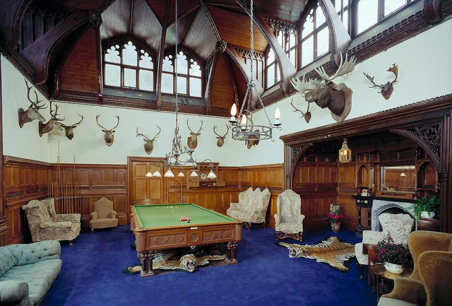 A billiards table stands in a tall hall with dark panelling, high windows and mounted antlers.