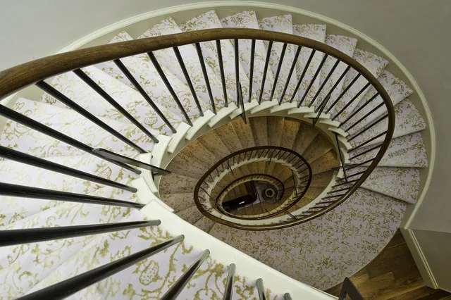 A view down an oval stairwell reveals patterned stair runners and dark railings winding through several floors.