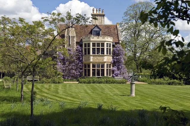 A tall projecting bay of windows overlooks a green lawn framed by flowering trees and leafy branches.
